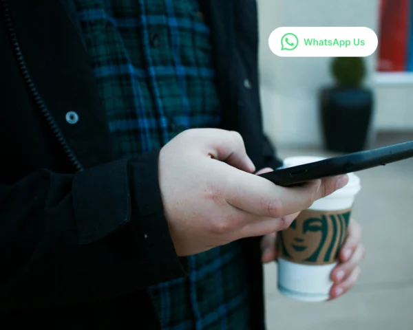 A man walking out of a coffee shop holding a takeaway coffee while chatting on WhatsApp with a Sandton plumbing company about fixing leaking bathroom pipes.